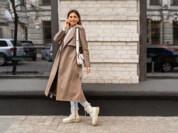 Jonge vrouw in lange beige trenchcoat met witte schoudertas, lichte jeans en beige boots